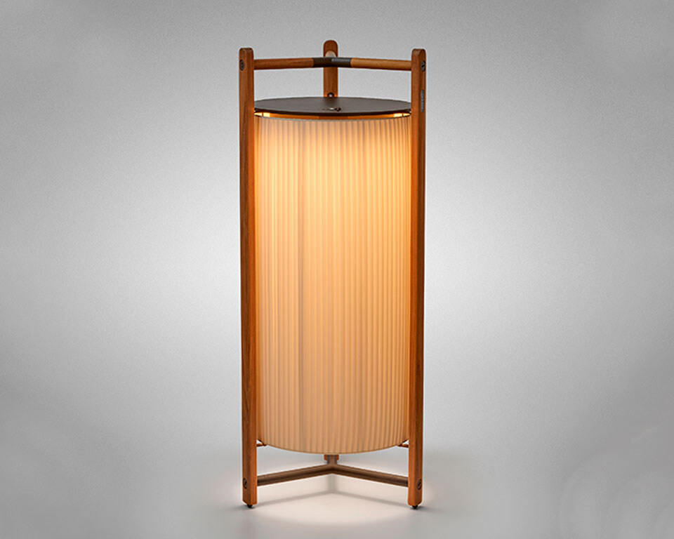 Kando-P111R-cream Eine moderne Tischlampe mit einer zylindrischen, geriffelten Schirmstruktur, umgeben von einem robusten dreibeinigen Rahmen aus Holz, der oben einen integrierten Griff aufweist. Die Lampe strahlt weiches Licht nach unten aus.