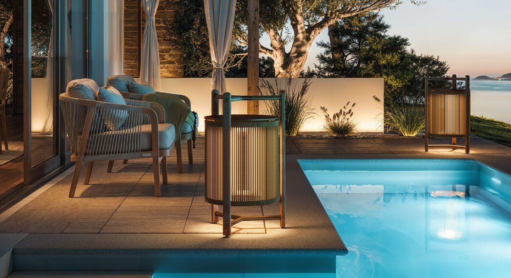 Kando P111R_Outdoor_1 Eine moderne Tischlampe mit einer zylindrischen, geriffelten Schirmstruktur, umgeben von einem robusten dreibeinigen Rahmen aus Holz, der oben einen integrierten Griff aufweist. Die Lampe strahlt weiches Licht nach unten aus.