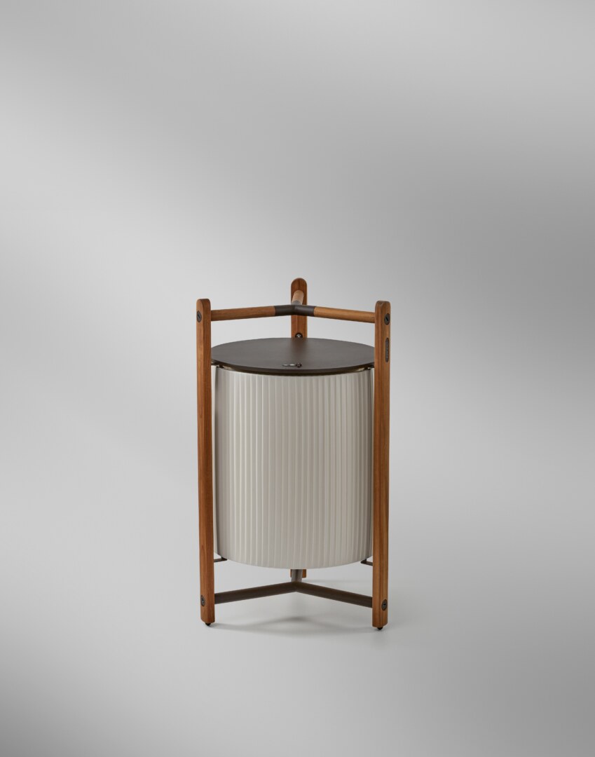 Kando P76R (6) Eine moderne Tischlampe mit einer zylindrischen, geriffelten Schirmstruktur, umgeben von einem robusten dreibeinigen Rahmen aus Holz, der oben einen integrierten Griff aufweist. Die Lampe strahlt weiches Licht nach unten aus.