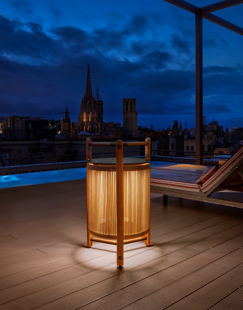 Kando P76R Outdoor Eine moderne Tischlampe mit einer zylindrischen, geriffelten Schirmstruktur, umgeben von einem robusten dreibeinigen Rahmen aus Holz, der oben einen integrierten Griff aufweist. Die Lampe strahlt weiches Licht nach unten aus.