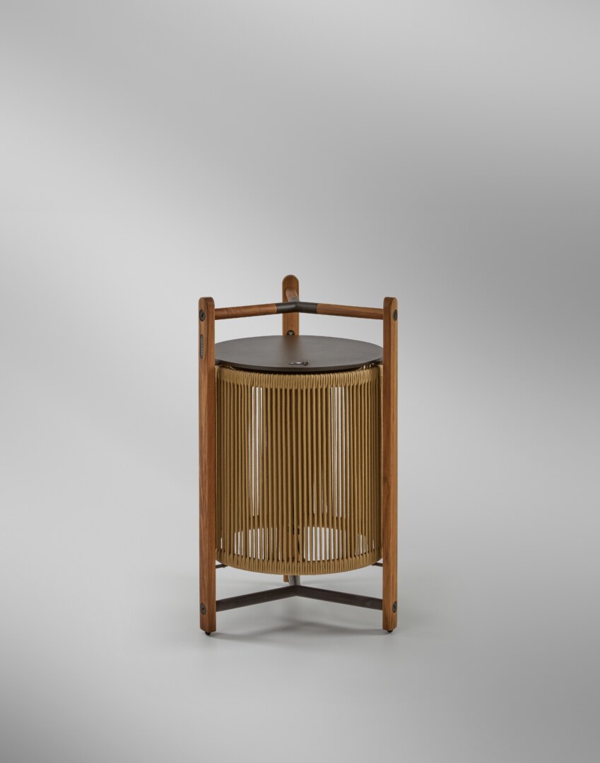 Kando P76R Outdoor Eine moderne Tischlampe mit einer zylindrischen, geriffelten Schirmstruktur, umgeben von einem robusten dreibeinigen Rahmen aus Holz, der oben einen integrierten Griff aufweist. Die Lampe strahlt weiches Licht nach unten aus.