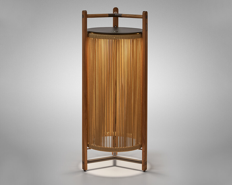 KandoP111R-Outdoor Eine moderne Tischlampe mit einer zylindrischen, geriffelten Schirmstruktur, umgeben von einem robusten dreibeinigen Rahmen aus Holz, der oben einen integrierten Griff aufweist. Die Lampe strahlt weiches Licht nach unten aus.