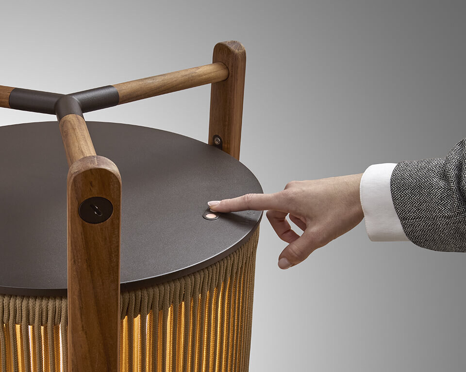 KandoP111R-Outdoor_B Eine moderne Tischlampe mit einer zylindrischen, geriffelten Schirmstruktur, umgeben von einem robusten dreibeinigen Rahmen aus Holz, der oben einen integrierten Griff aufweist. Die Lampe strahlt weiches Licht nach unten aus.