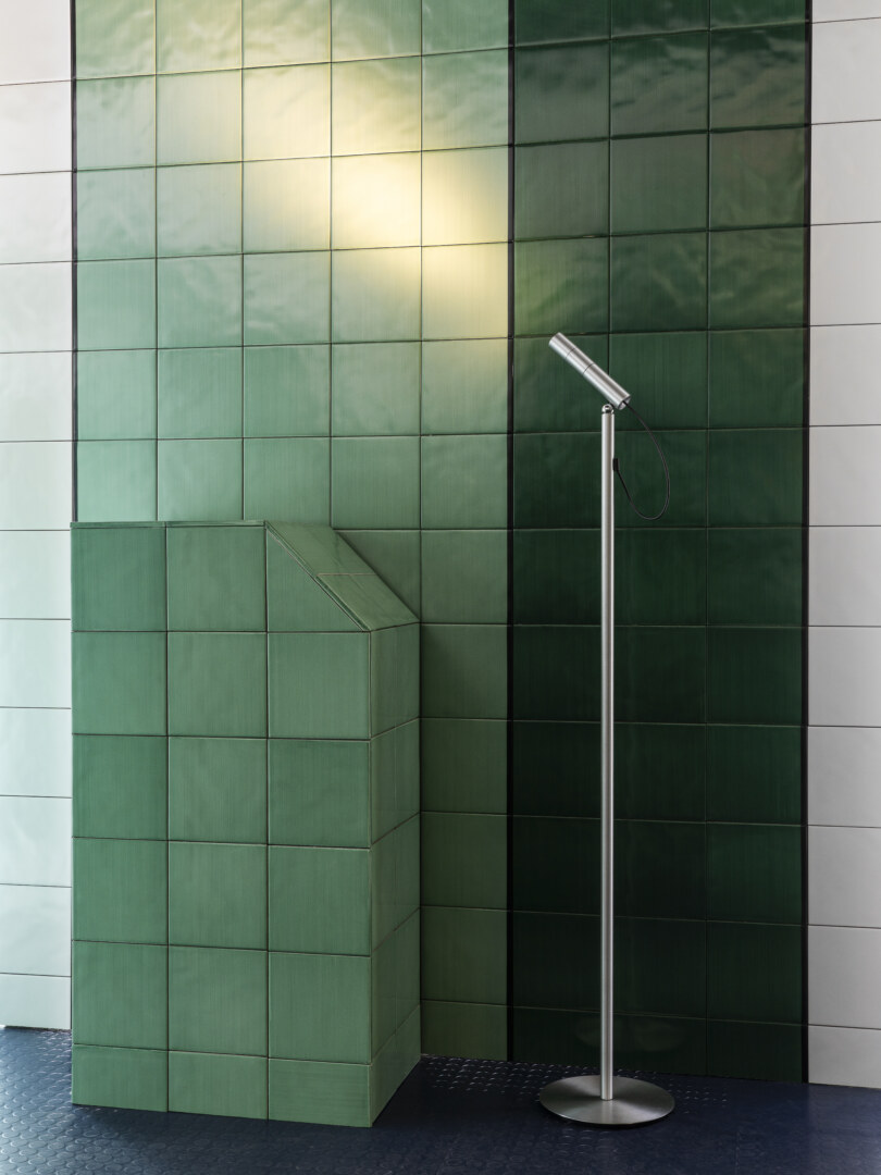 Eine Stehlampe aus Edelstahl steht in einem Raum, der mit grün- und weiss-glasierten Kacheln verkleidet ist. Die Lampe wirft ein warmes Licht auf die grünen Fliesen.