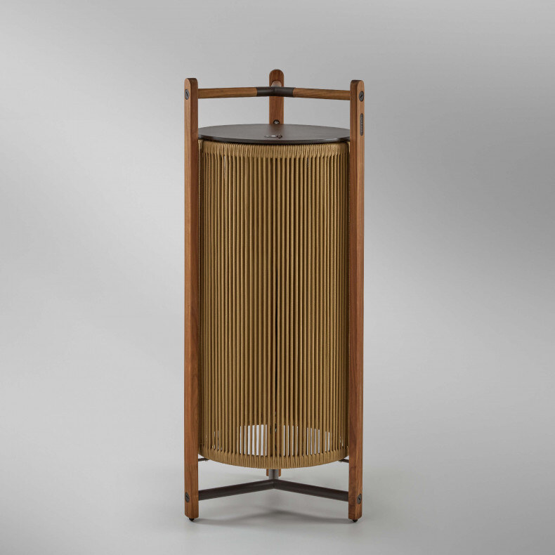 kando-p111r-outdoor Eine moderne Tischlampe mit einer zylindrischen, geriffelten Schirmstruktur, umgeben von einem robusten dreibeinigen Rahmen aus Holz, der oben einen integrierten Griff aufweist. Die Lampe strahlt weiches Licht nach unten aus.