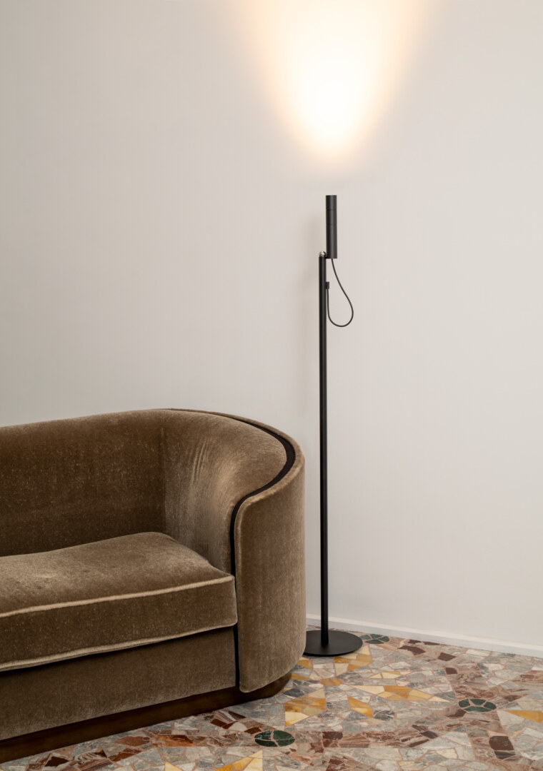 Eine moderne Stehlampe steht neben einem beigen Sofa in einem stilvoll eingerichteten Raum mit einem Mosaikboden. Die Lampe strahlt ein warmes Licht an die weisse Wand.