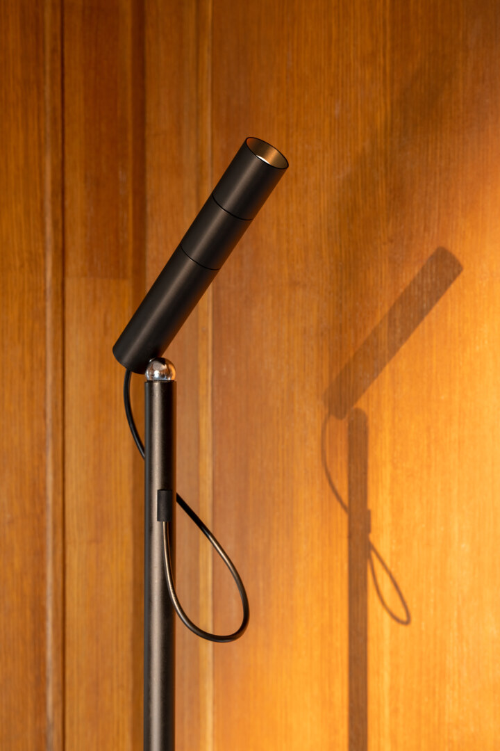 Ein Nahaufnahmebild einer schwarzen Stehlampe vor einer Holzverkleidung. Der zylinderförmige Leuchtkopf und die minimalistische Bauweise der Lampe sind deutlich zu erkennen.