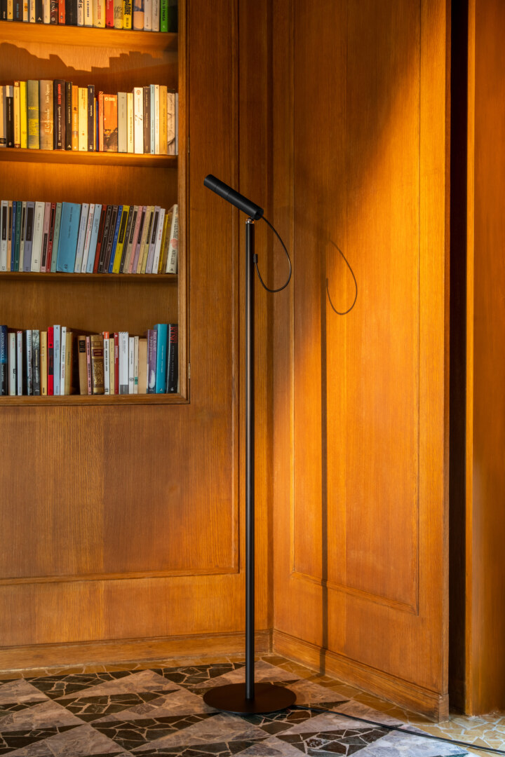 Eine schwarze Stehlampe steht in einer gemütlichen Bibliothek neben einem Holzregal, das mit Büchern gefüllt ist. Das warme Licht der Lampe beleuchtet die Holzwände.